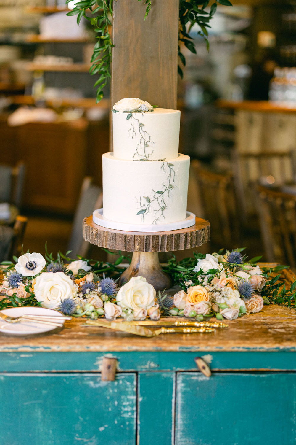 A beautifully arranged three-tier wedding cake adorned with floral accents, sitting atop a wooden cake stand, surrounded by an array of fresh flowers on a rustic table.