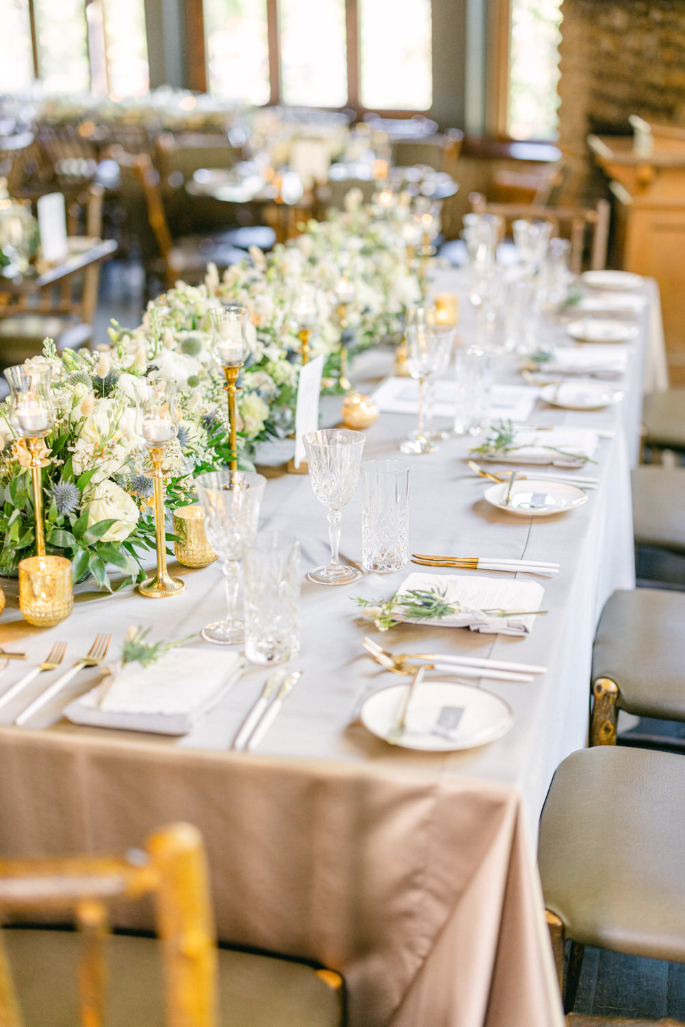 A beautifully arranged dining table with floral centerpieces, glassware, and gold accents set for a wedding reception.