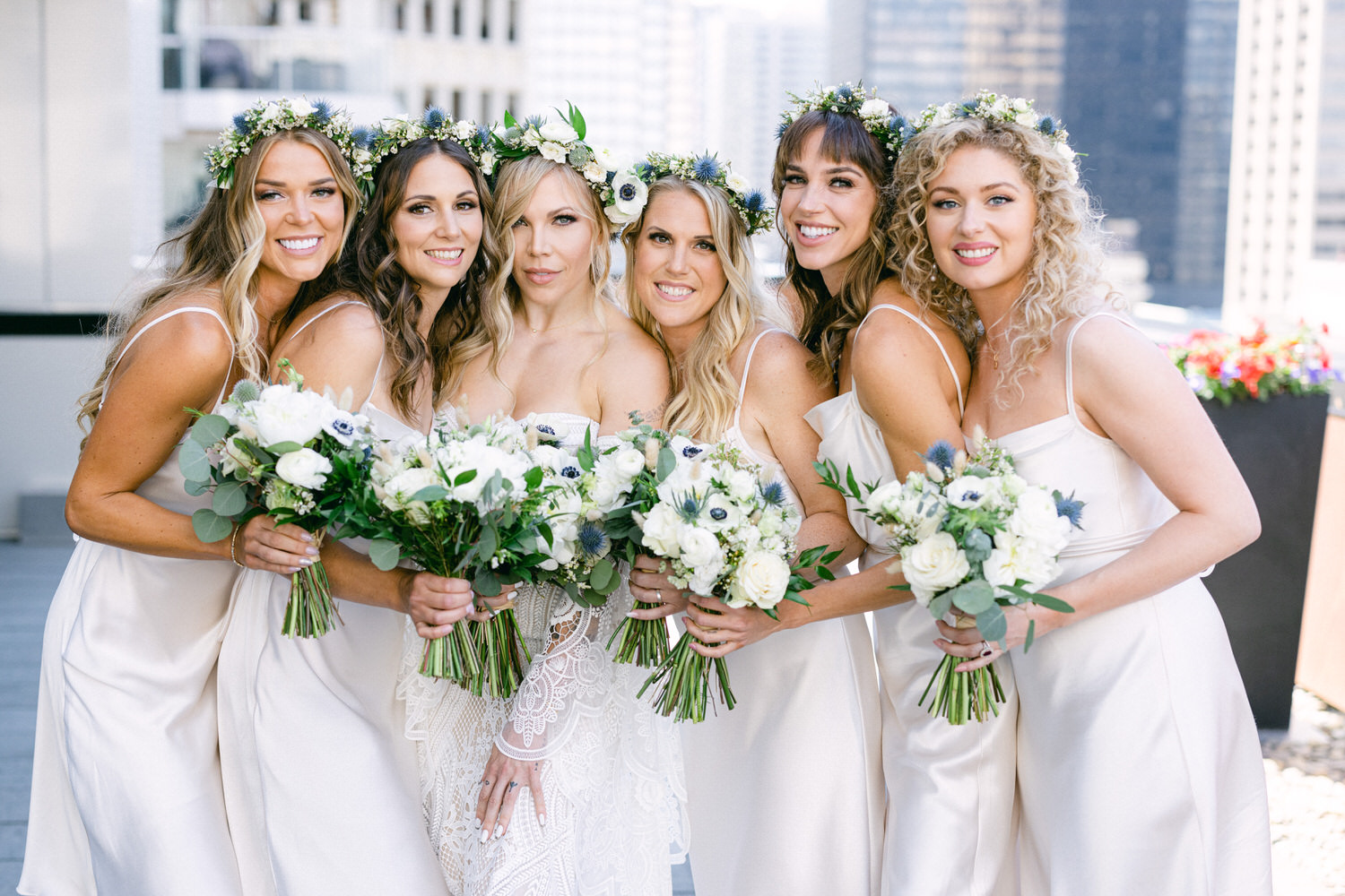 A joyful group of six bridesmaids wearing elegant satin dresses and floral crowns, holding bouquets of white and green flowers.