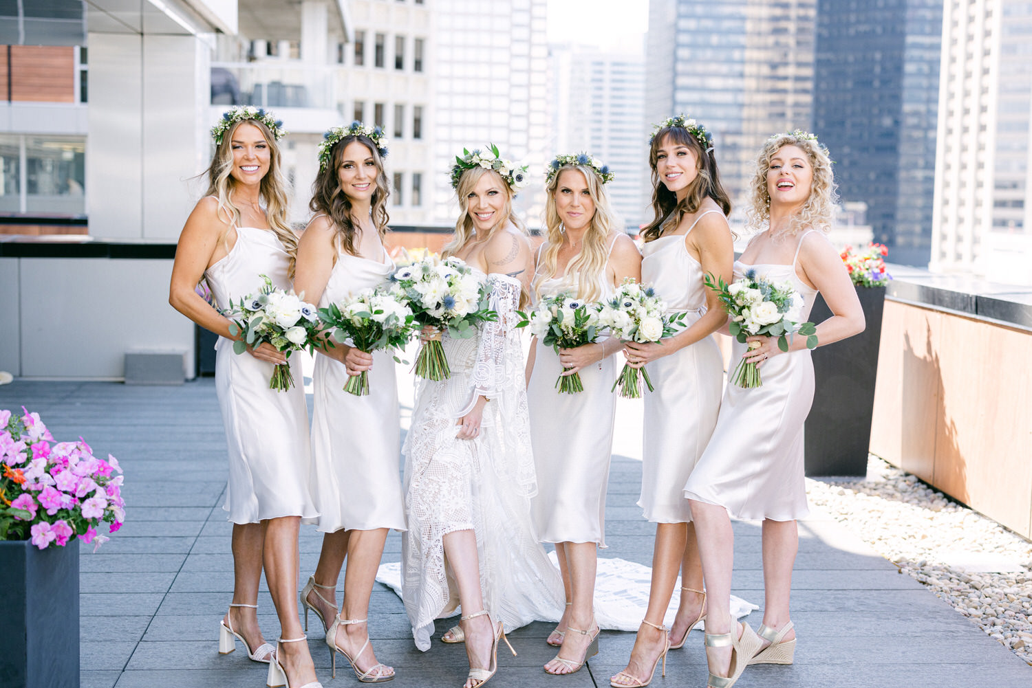 A joyful bridal party poses on a rooftop, wearing elegant satin dresses and floral crowns, each holding bouquets of white flowers.