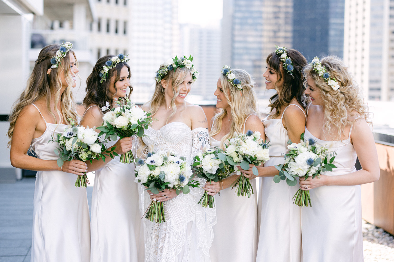 A joyful bridal party of six women in elegant satin dresses, adorned with floral crowns, smiling and holding white floral bouquets against a city skyline backdrop.