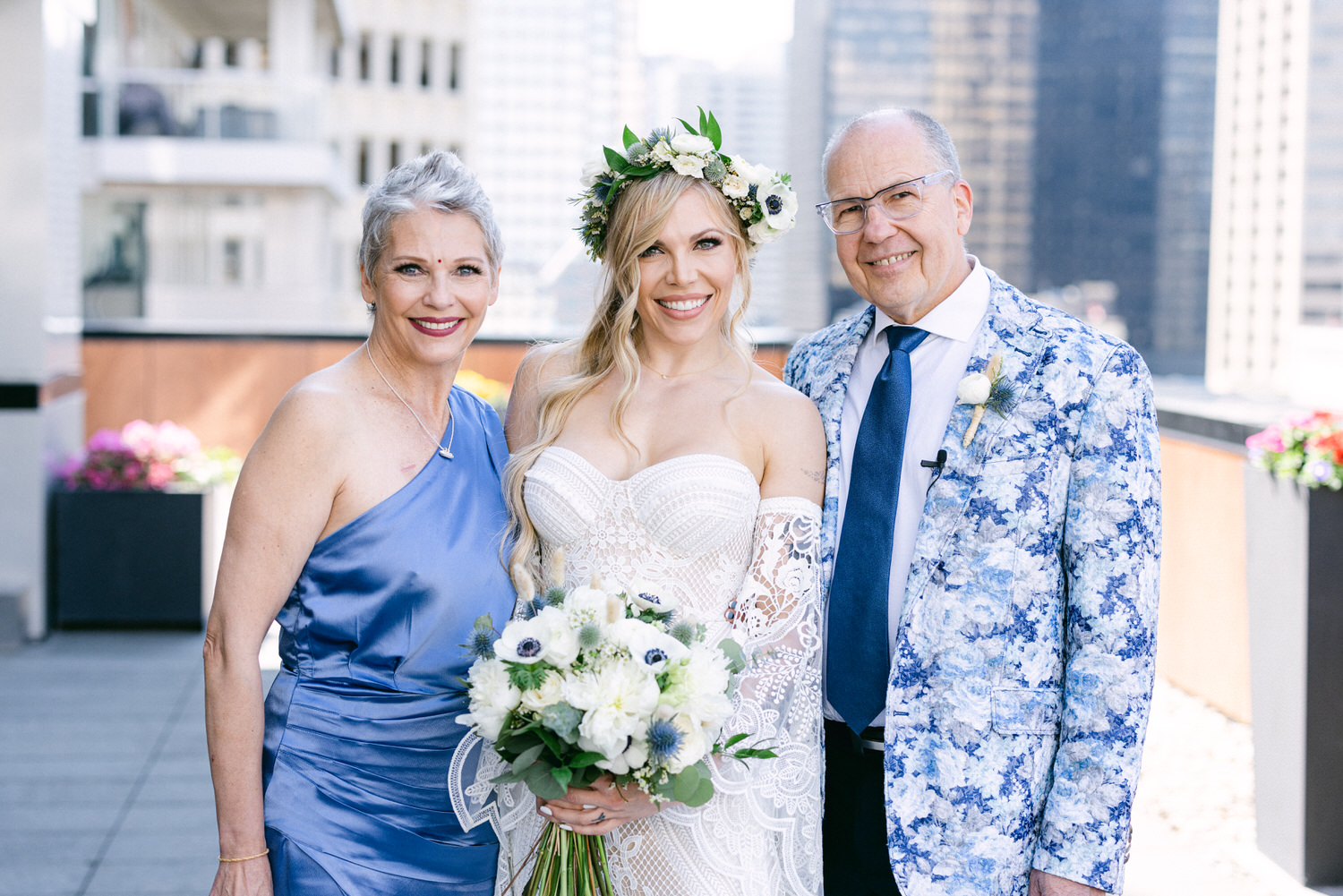 A bride stands with her parents, all smiling, against a backdrop of a city skyline, showcasing floral decorations and outfits that blend elegance with vibrant colors.