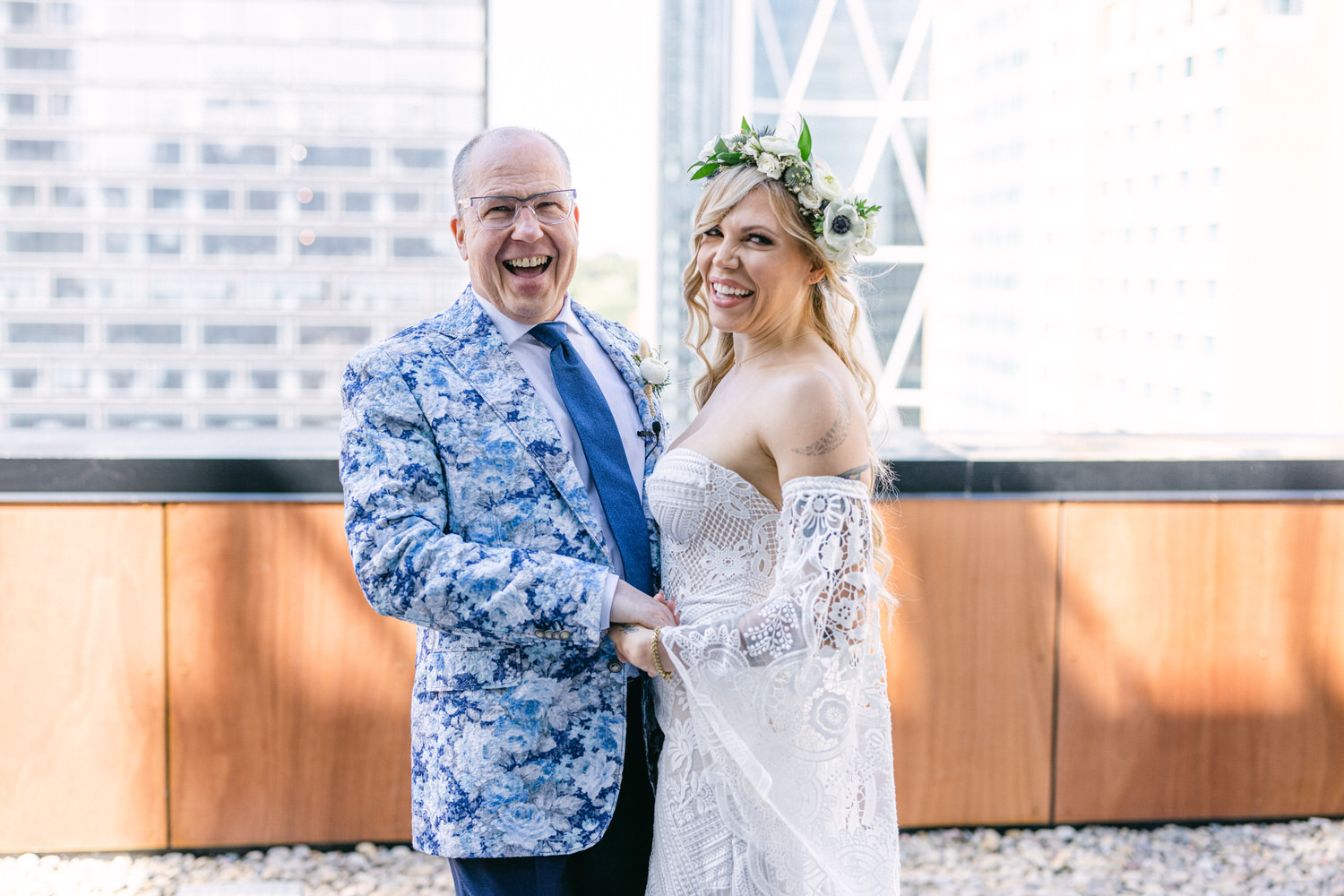 A smiling couple shares a joyful moment outdoors, with the man wearing a blue floral suit and the woman in a lace wedding gown adorned with a flower crown.