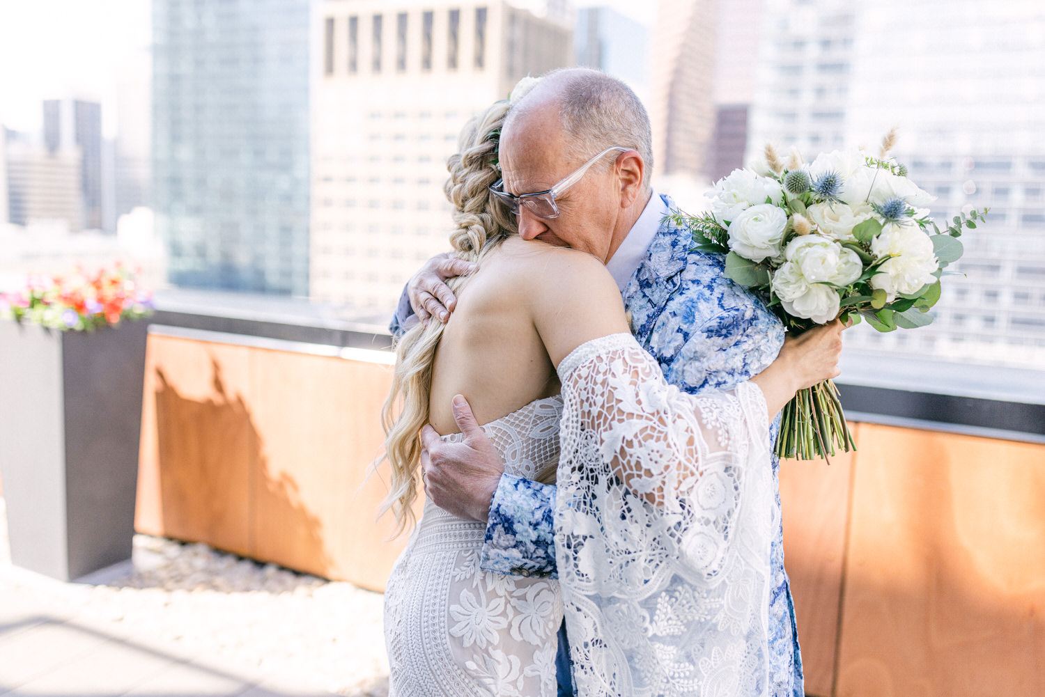 A bride and her father share a heartfelt hug, with the city skyline in the background and the bride holding a bouquet of flowers.