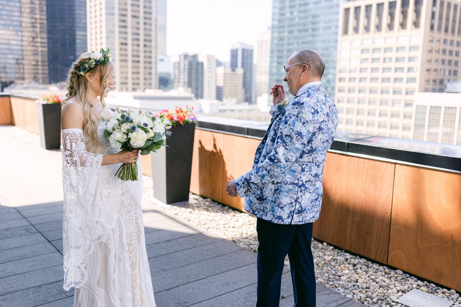 A bride in a lace gown and floral crown smiles at a man in a patterned suit, surrounded by a modern cityscape.