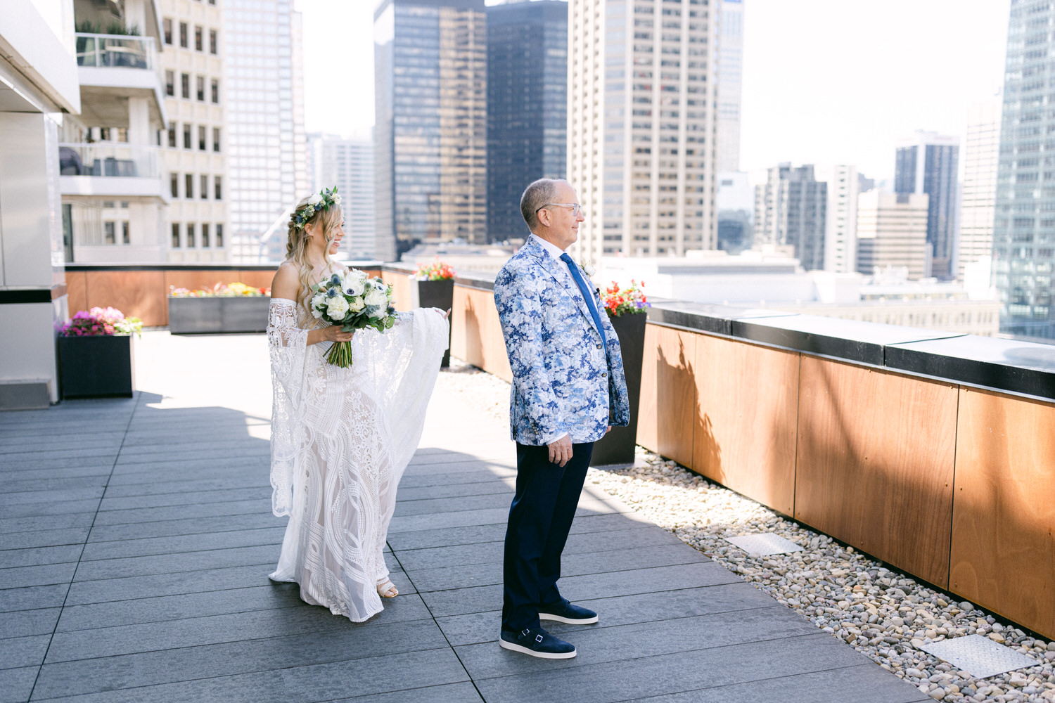 Bride in a flowing lace gown holding a bouquet approaches a man in a patterned blazer on a city rooftop.