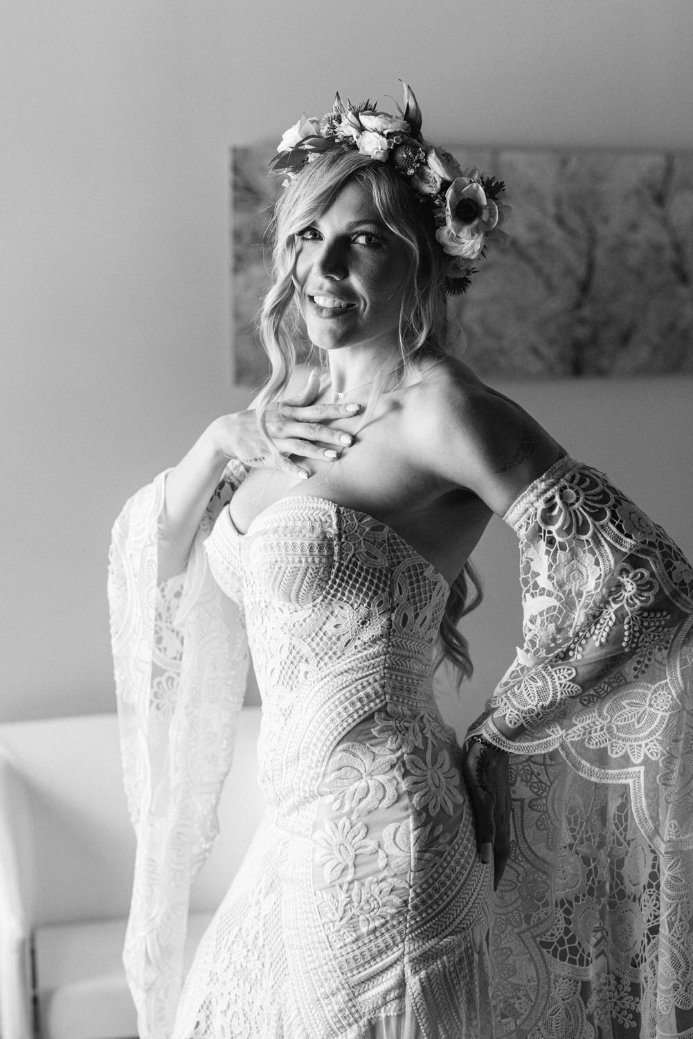 A joyful bride showcasing her intricate lace gown and floral crown, striking a playful pose with a warm smile.