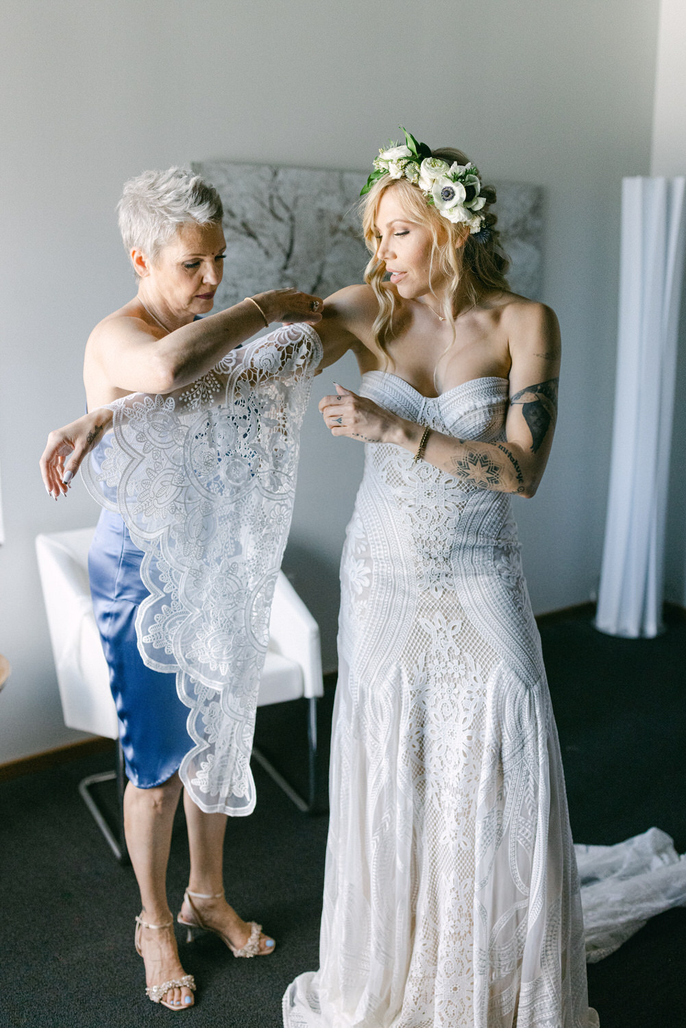 A bride in a fitted lace wedding dress stands beside her mother, who is helping her put on an elegant lace shawl, in a bright and modern setting.