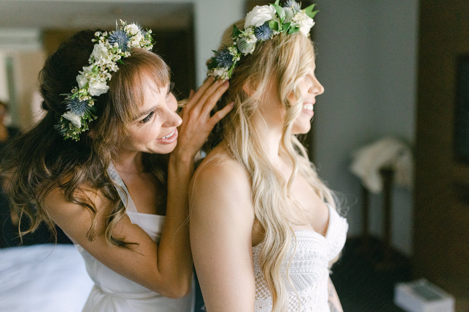 Two bridesmaids share a joyful moment while adjusting each other's floral crowns in a softly lit setting.