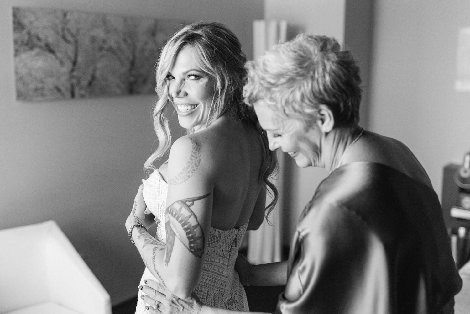 A bride smiles happily while her mother assists her with getting ready, showcasing an intimate and joyful bond between them.