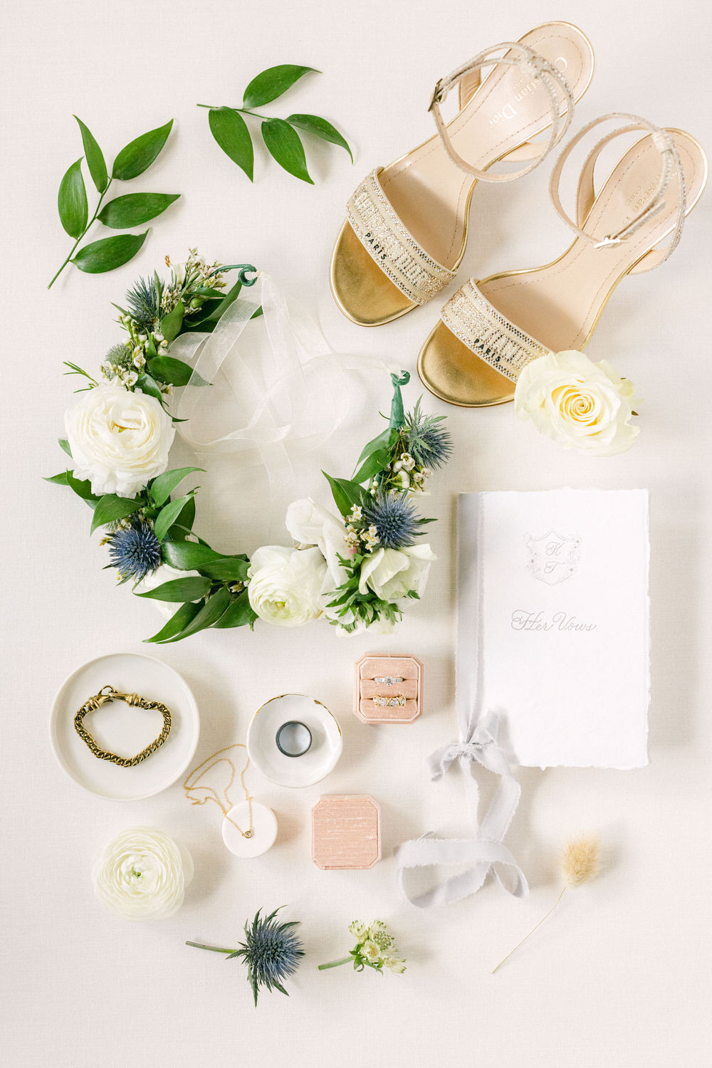 Bridal Accessories and Keepsakes:::A flat lay arrangement featuring elegant bridal sandals, a floral crown, vow book, and various jewelry pieces against a soft background.