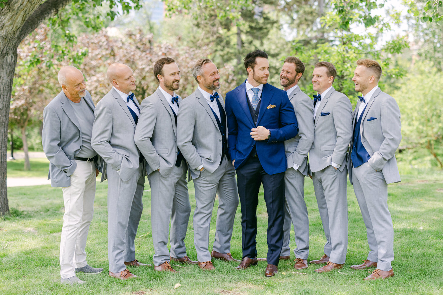 A group of well-dressed men in suits gather outdoors, with the groom in a blue suit at the center, smiling and interacting with his groomsmen in gray suits.