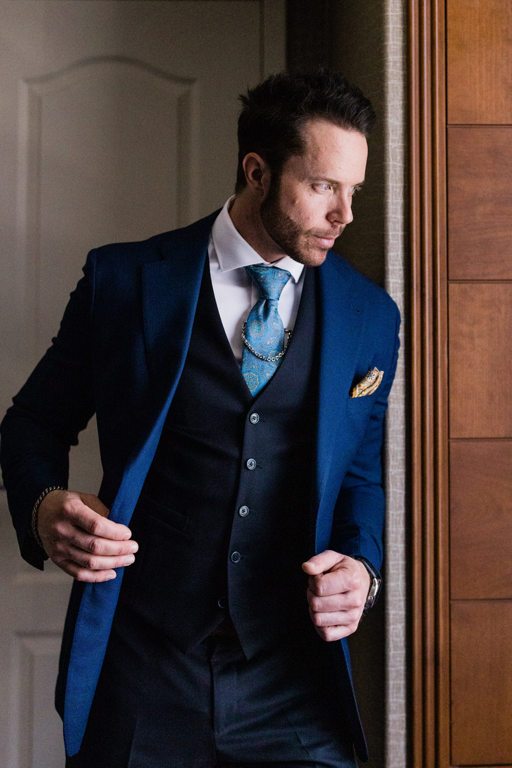 A man dressed in a stylish blue suit with a patterned tie and pocket square, standing thoughtfully by a window.