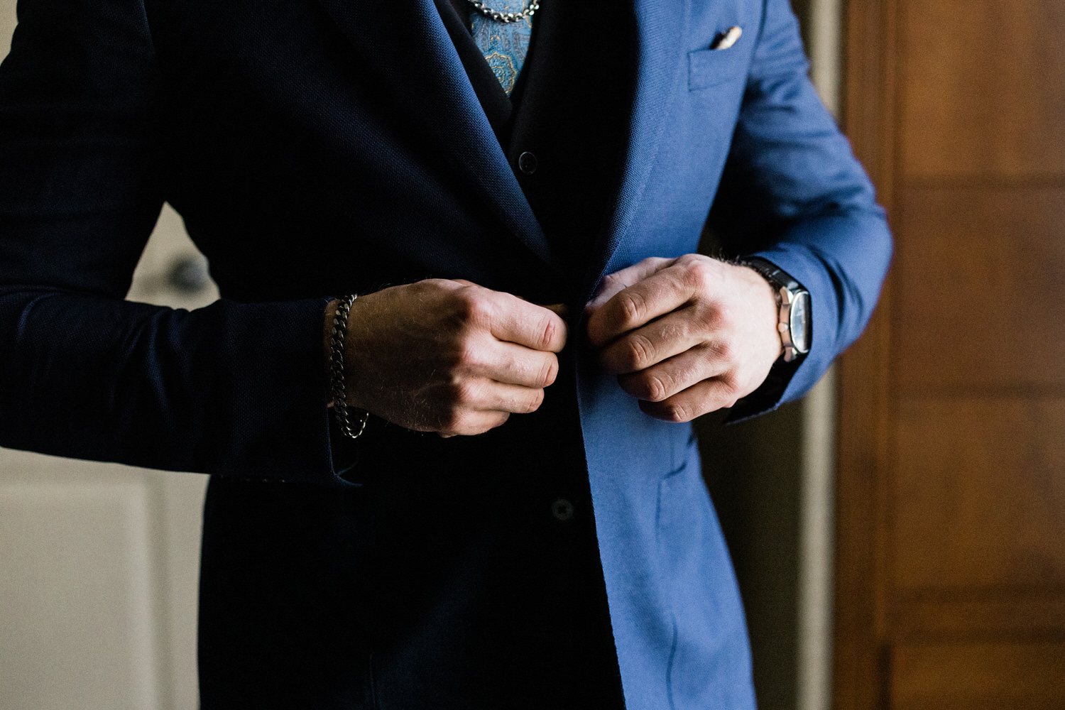 Close-up of a man's hands buttoning a blue suit jacket, showcasing stylish accessories and elegant attire.