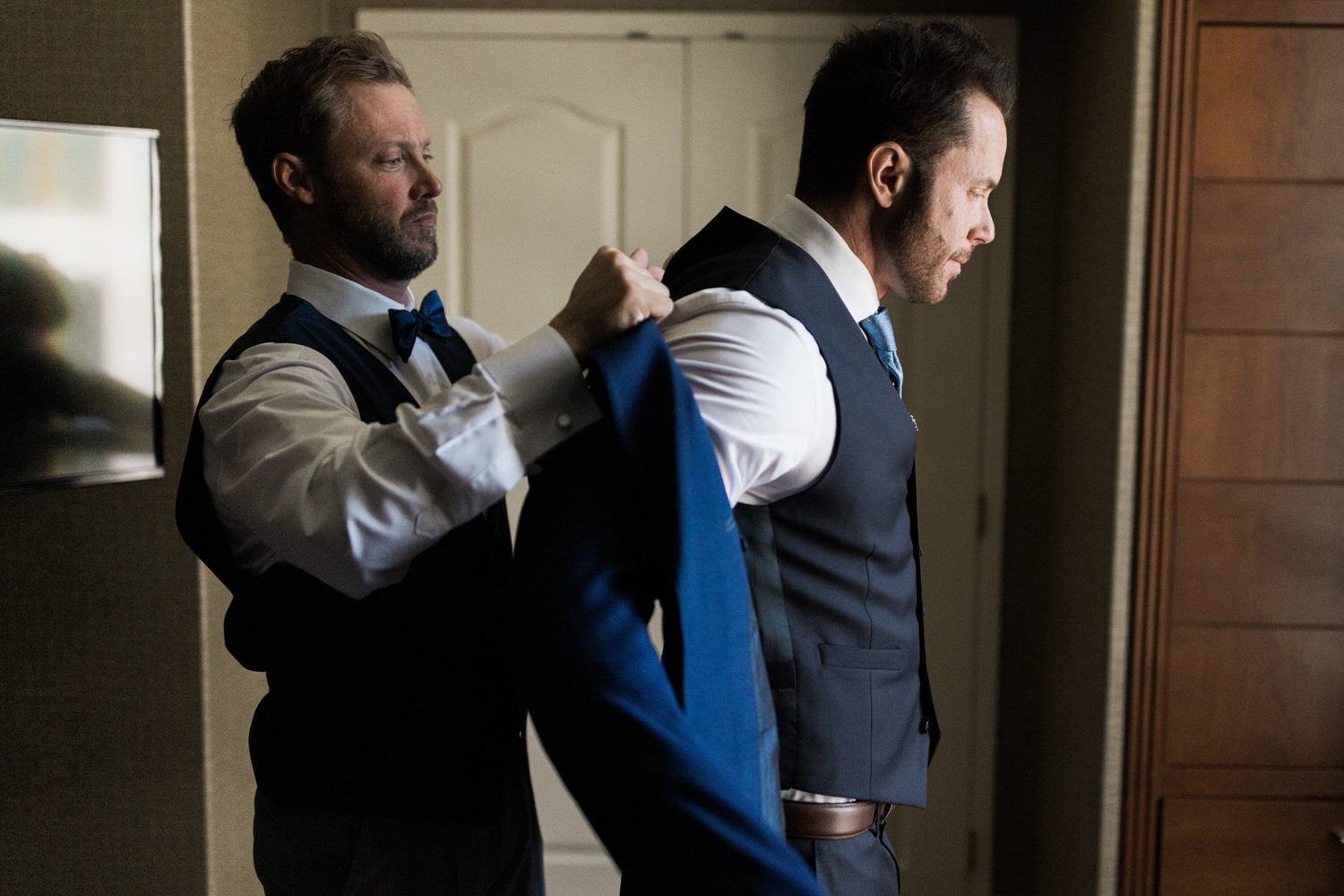 A man assists another in putting on a formal suit in a well-lit room, showing moments of preparation before a wedding.