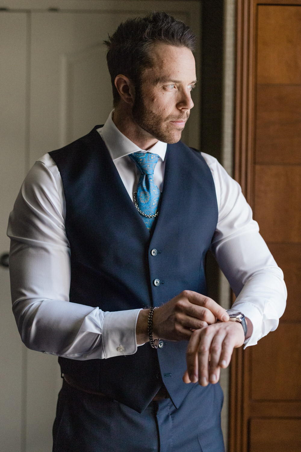 Man in formal attire adjusts his watch while looking thoughtfully out a window, exuding elegance and anticipation.