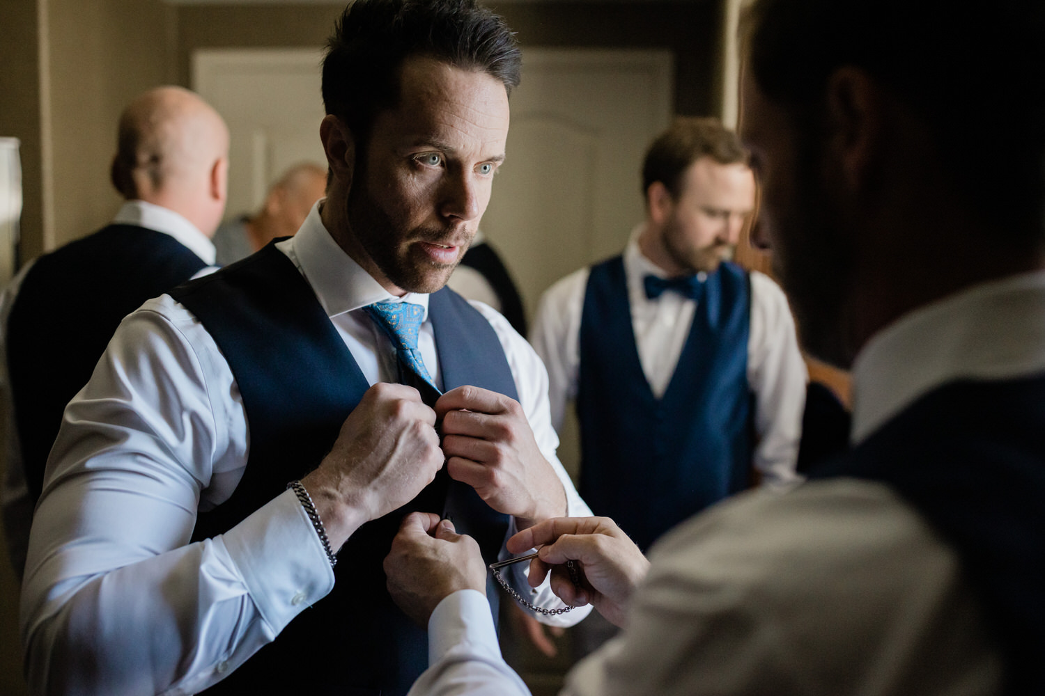 A groom adjusts his tie with the help of a friend while other groomsmen prepare in the background.