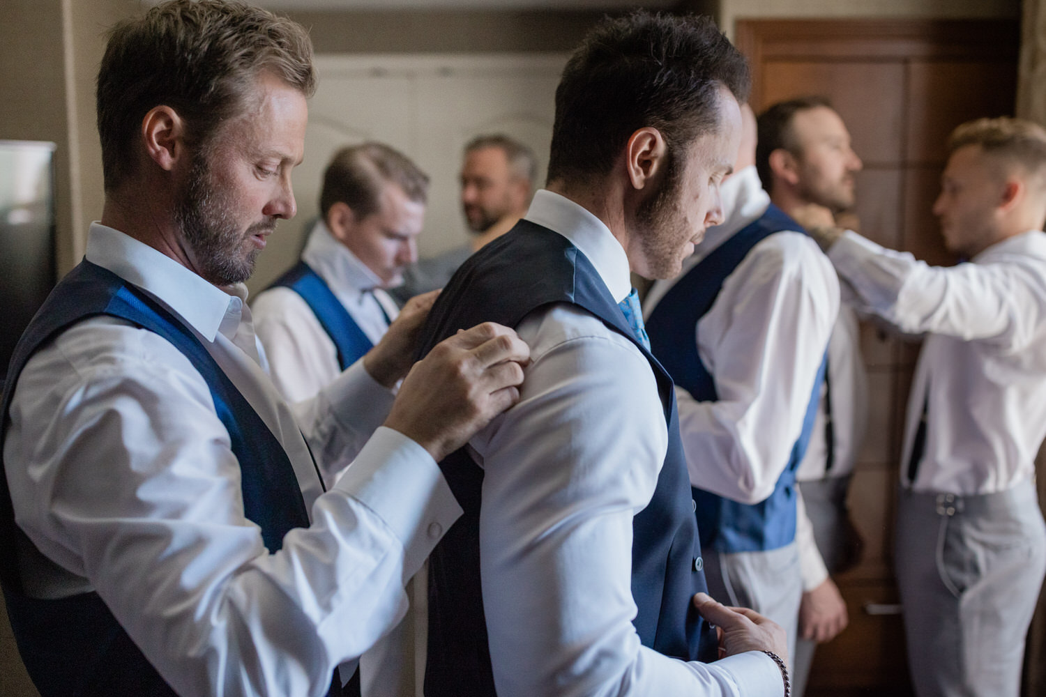 A group of men adjusting their formal attire, preparing for a wedding ceremony, with focus on a groom having his vest fitted.