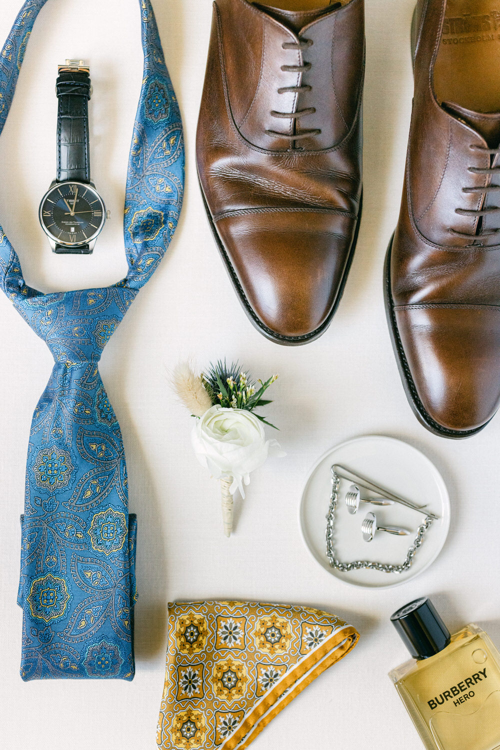 An arrangement of a blue paisley tie, elegant brown dress shoes, a black wristwatch, boutonniere, yellow patterned pocket square, silver cufflinks, and Burberry perfume on a white background.