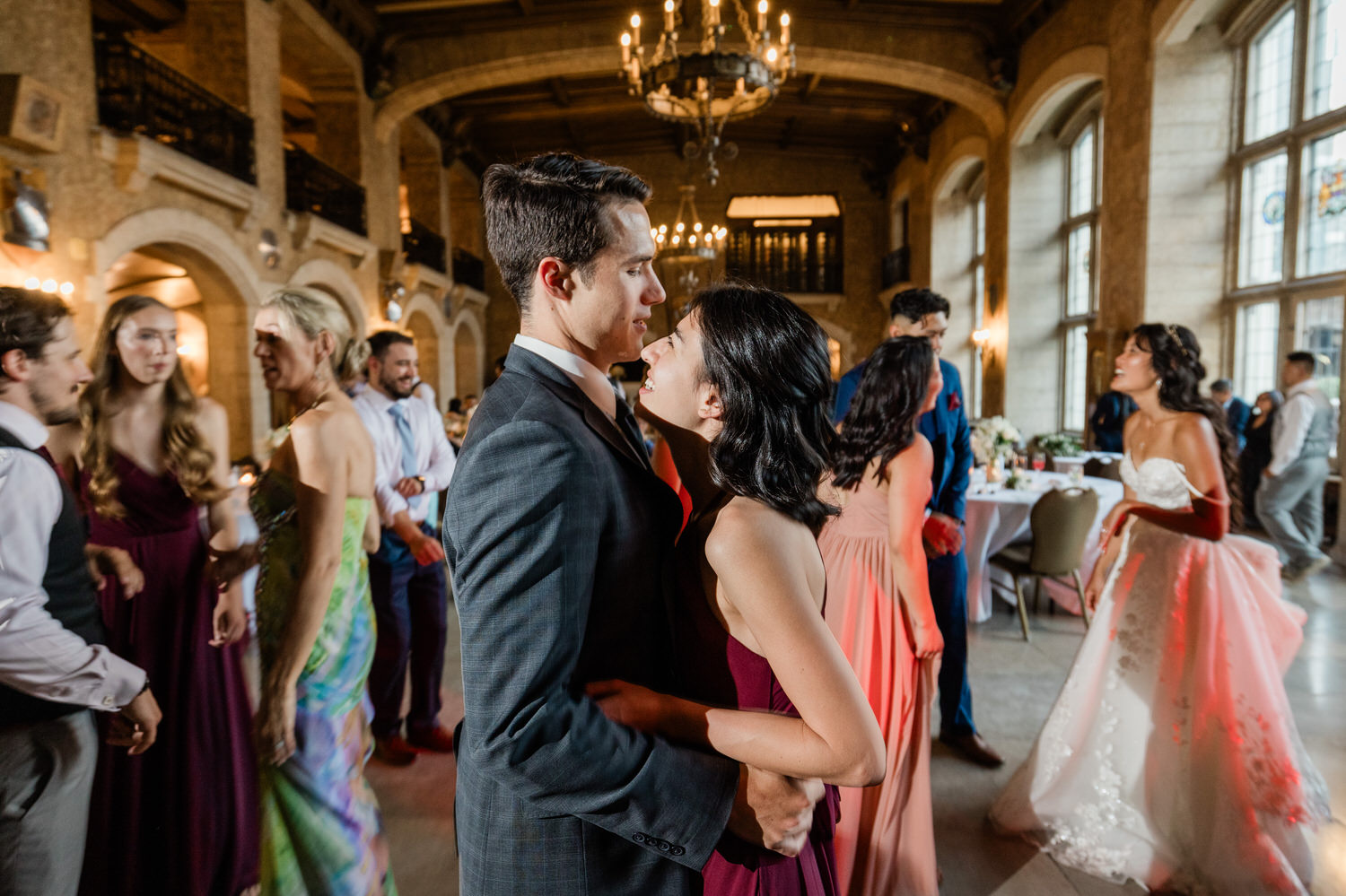 A couple dances closely at a vibrant wedding reception, surrounded by guests in elegant attire enjoying the celebration in an ornate venue.