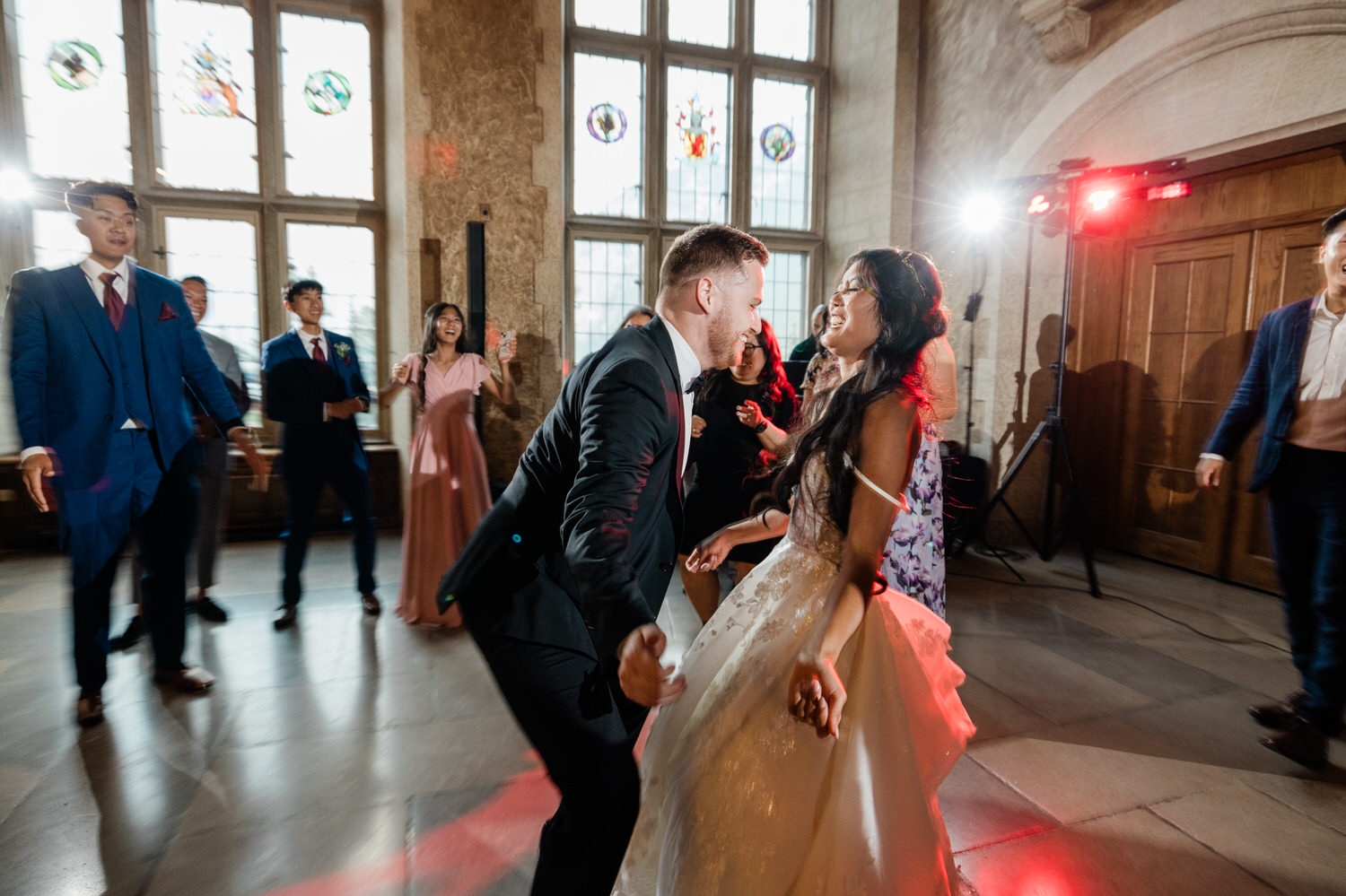 Joyful moment of a couple dancing amidst guests at a wedding reception, with vibrant lighting and a festive atmosphere.