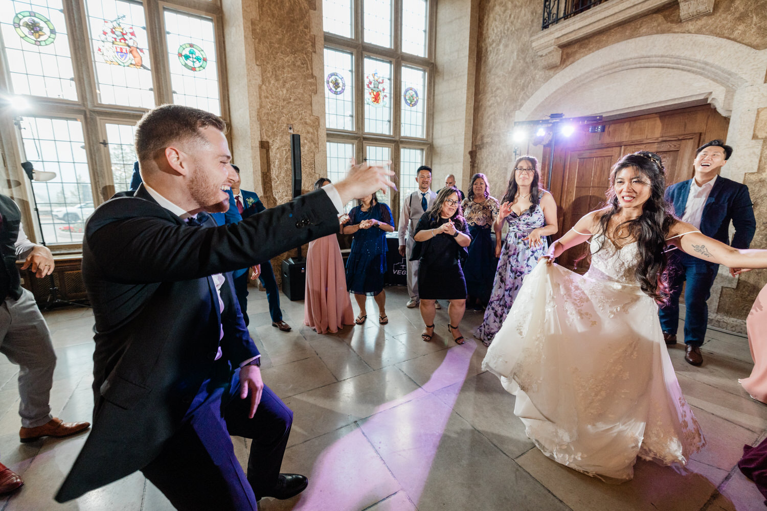 A joyful scene of wedding guests dancing, featuring a bride in a flowing dress and a groom engaging with the crowd in a stunning venue with stained glass windows.