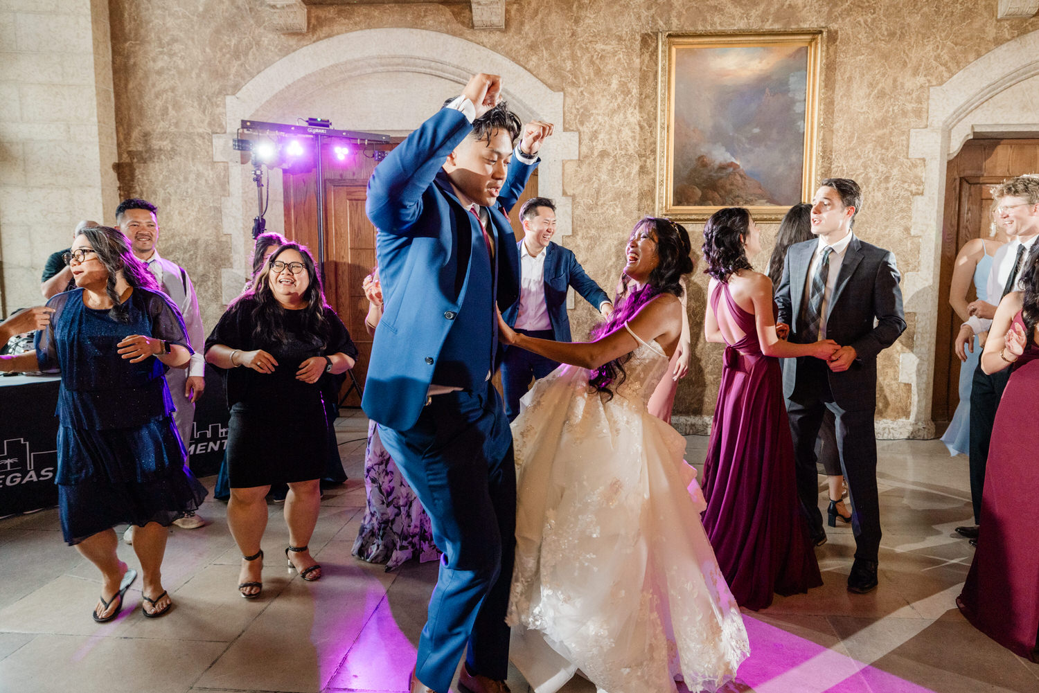 A lively group of guests dancing joyfully at a wedding reception, with a blend of formal attire and vibrant lighting in an elegant venue.