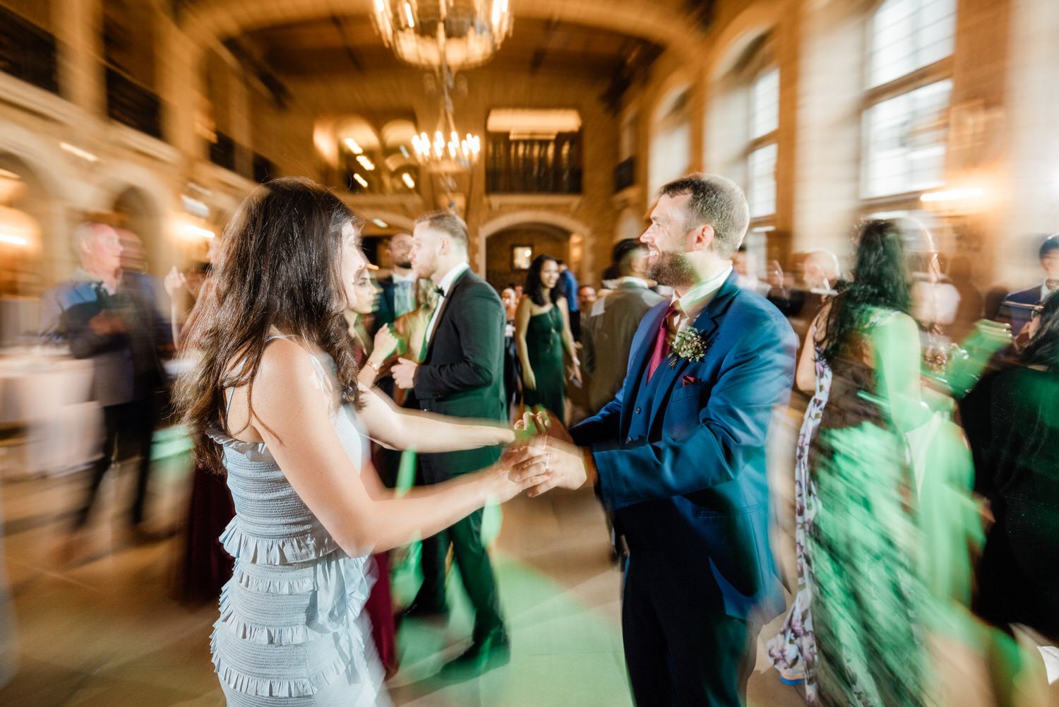 A couple dances joyfully amidst a lively wedding reception, surrounded by guests in elegant attire, with a beautiful venue backdrop and dynamic movement capturing the festive atmosphere.