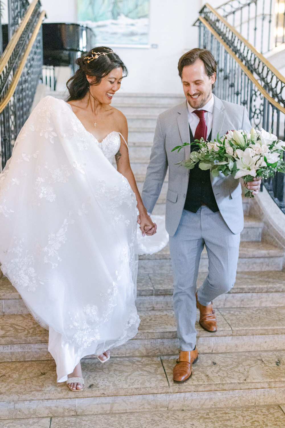 A bride in a flowing white gown and a groom in a light gray suit hold hands and walk down a staircase, smiling at each other amidst elegant surroundings.