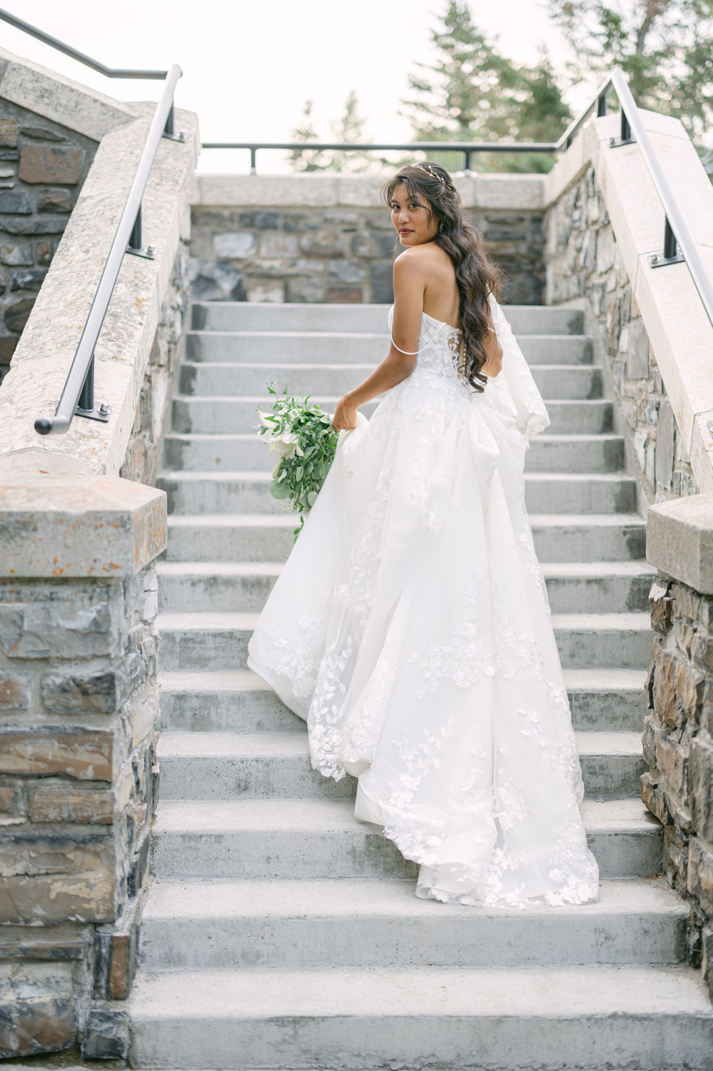 A bride in a stunning white gown with intricate floral embroidery, holding a bouquet, gracefully walks up stone steps surrounded by greenery.