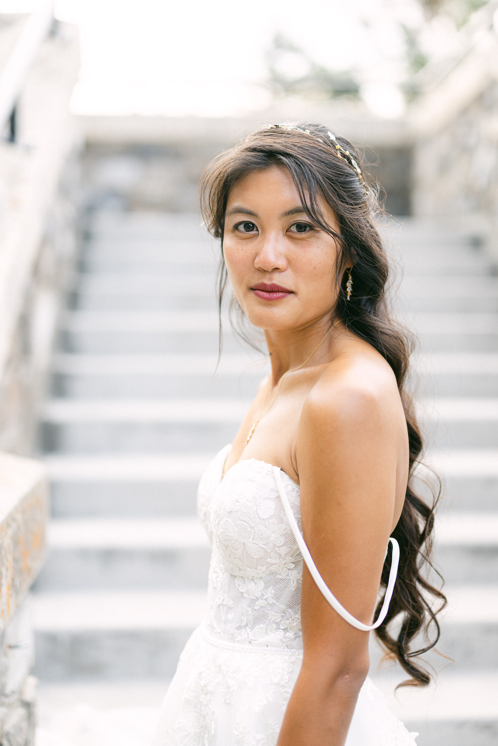 A bride in a stunning white gown gazes elegantly at the camera, with soft waves in her hair and a delicate hair accessory, standing on an outdoor staircase.