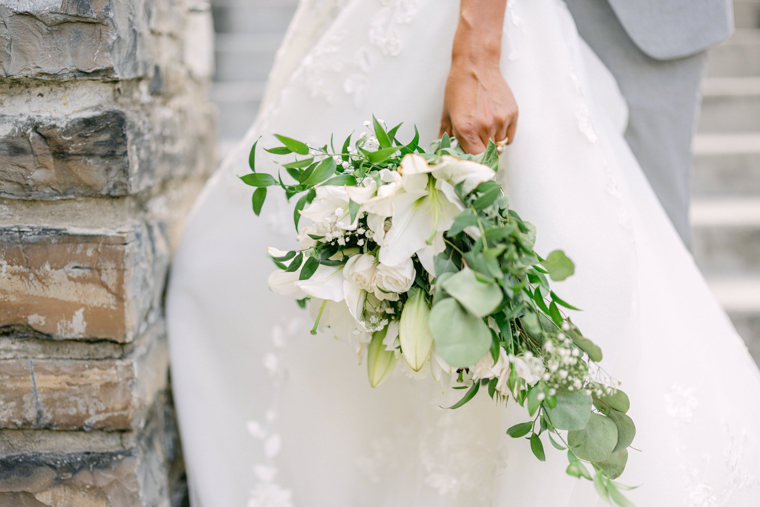 Close-up of a bride's hand holding a bouquet of white flowers and greenery, with a wedding dress partially visible and stone wall in the background.