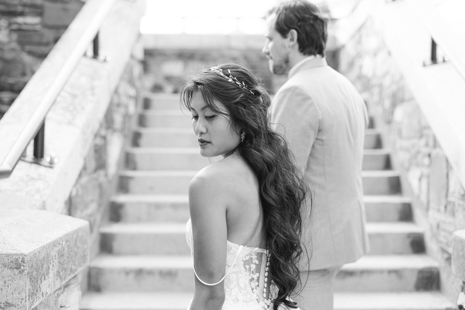 A bride with long hair in a beautiful gown looks thoughtfully over her shoulder, while a groom stands behind her on the stairs.