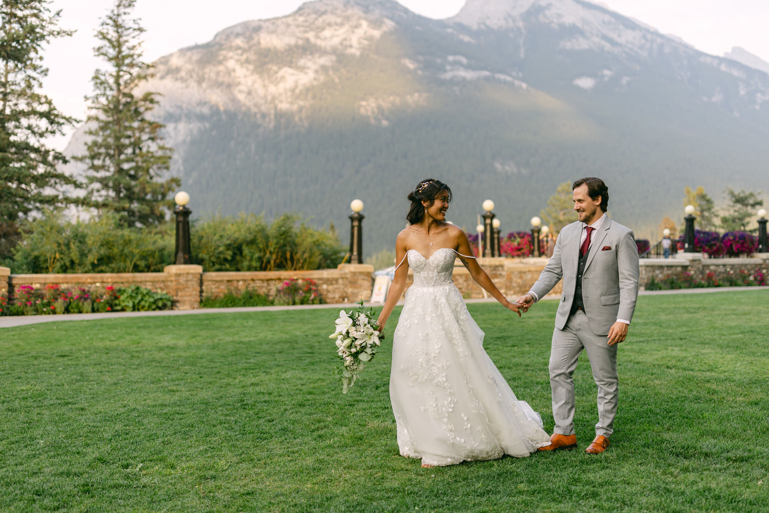 A couple walks hand-in-hand on a lush green lawn, surrounded by majestic mountains and vibrant flowers, capturing a moment of joy and love on their special day.