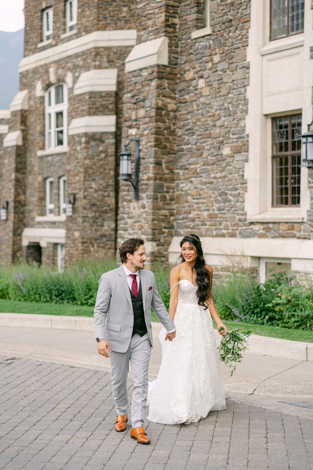 A bride in a flowing white gown and a groom in a light gray suit walk hand-in-hand along a stone pathway, surrounded by lush greenery and an elegant stone building in the background.