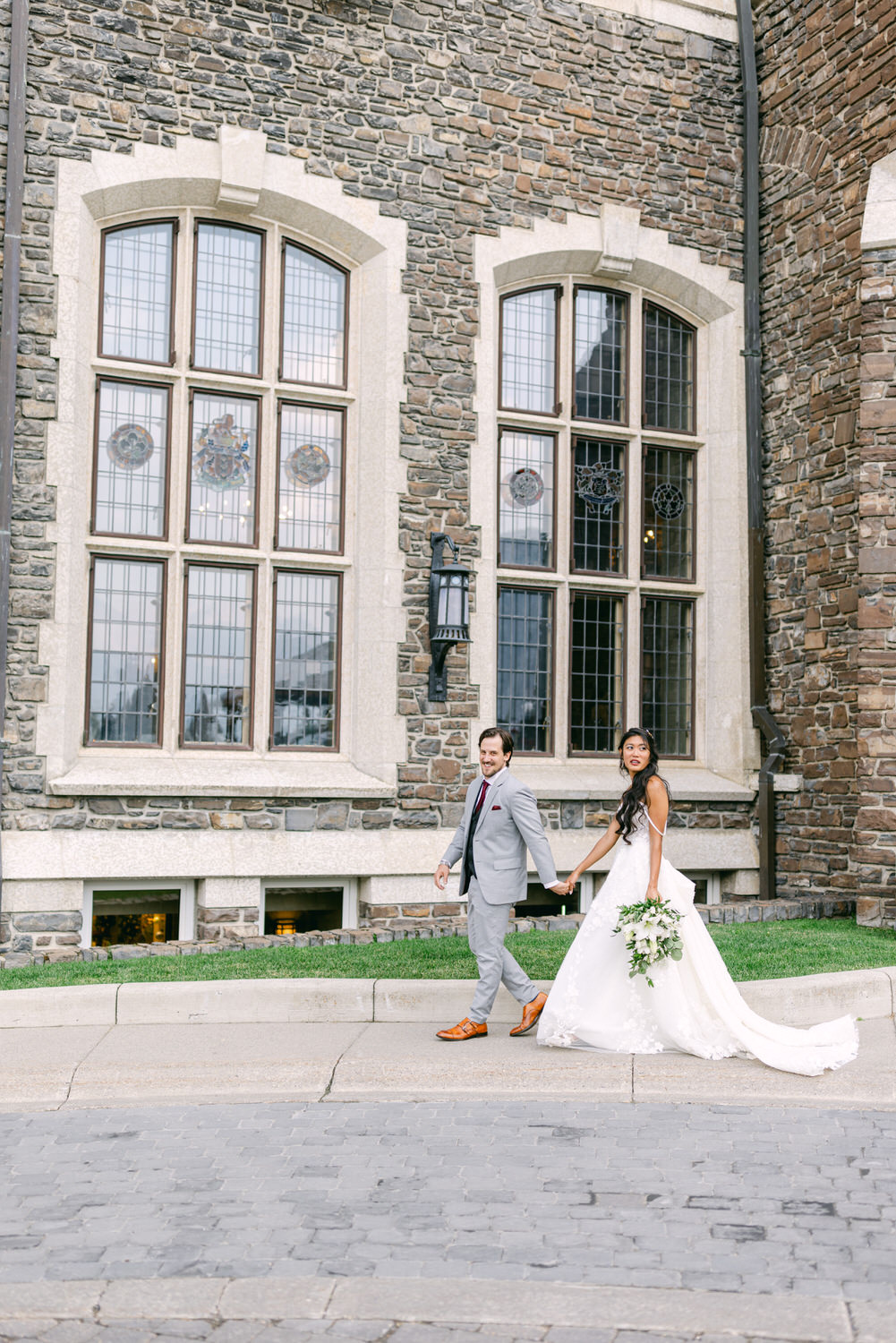 A joyful couple walks hand in hand, dressed in wedding attire, in front of a charming stone building with large windows and intricate stained glass.