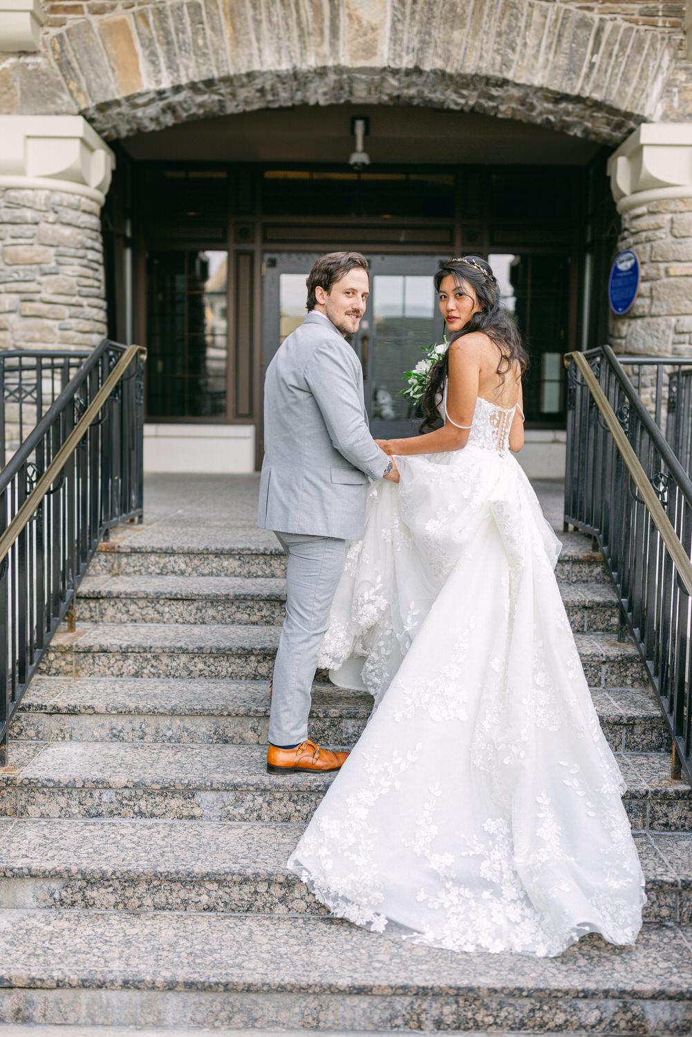 A bride and groom pose together on stone steps, with the bride looking back over her shoulder, showcasing her elegant wedding dress and bouquet while the groom holds her hand.