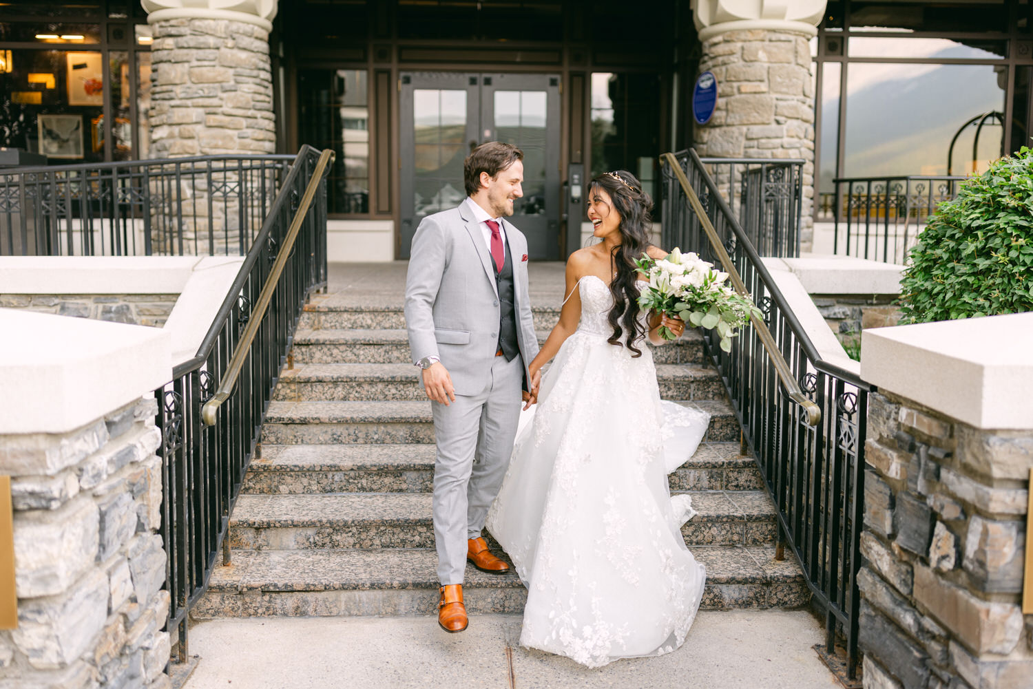 A happy couple walks hand in hand down stone steps, with the bride in a flowing white gown and the groom in a light gray suit, amidst a scenic backdrop.