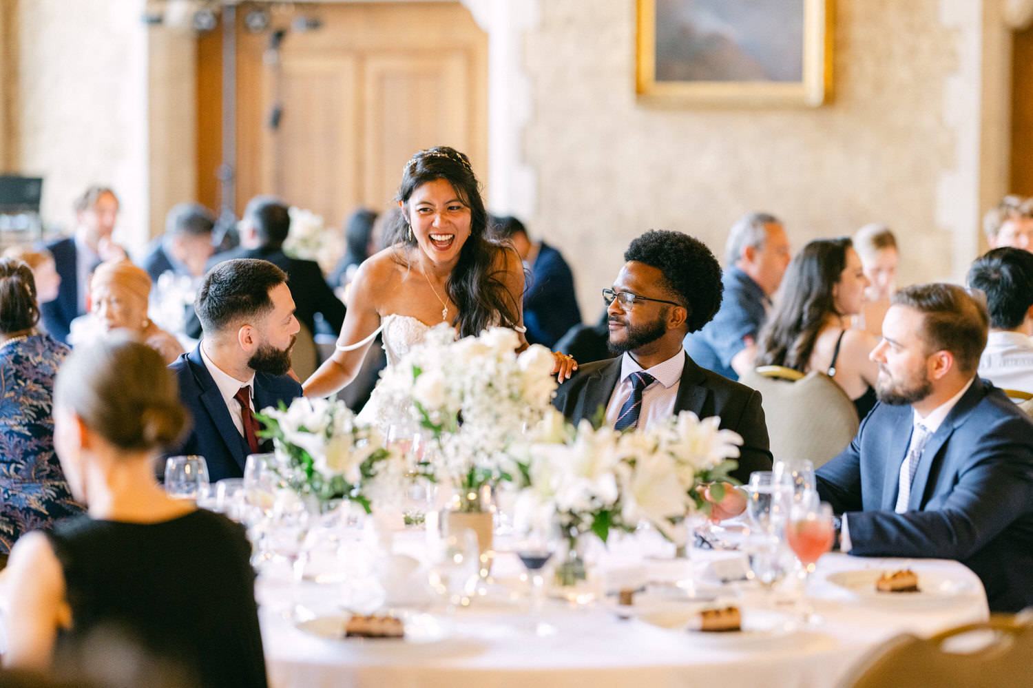 A bride joyfully interacts with guests at a wedding reception, surrounded by floral arrangements and elegantly set tables.