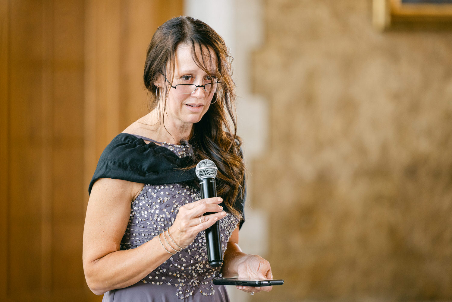 A woman in a beaded purple dress holds a microphone and smartphone while giving a speech.