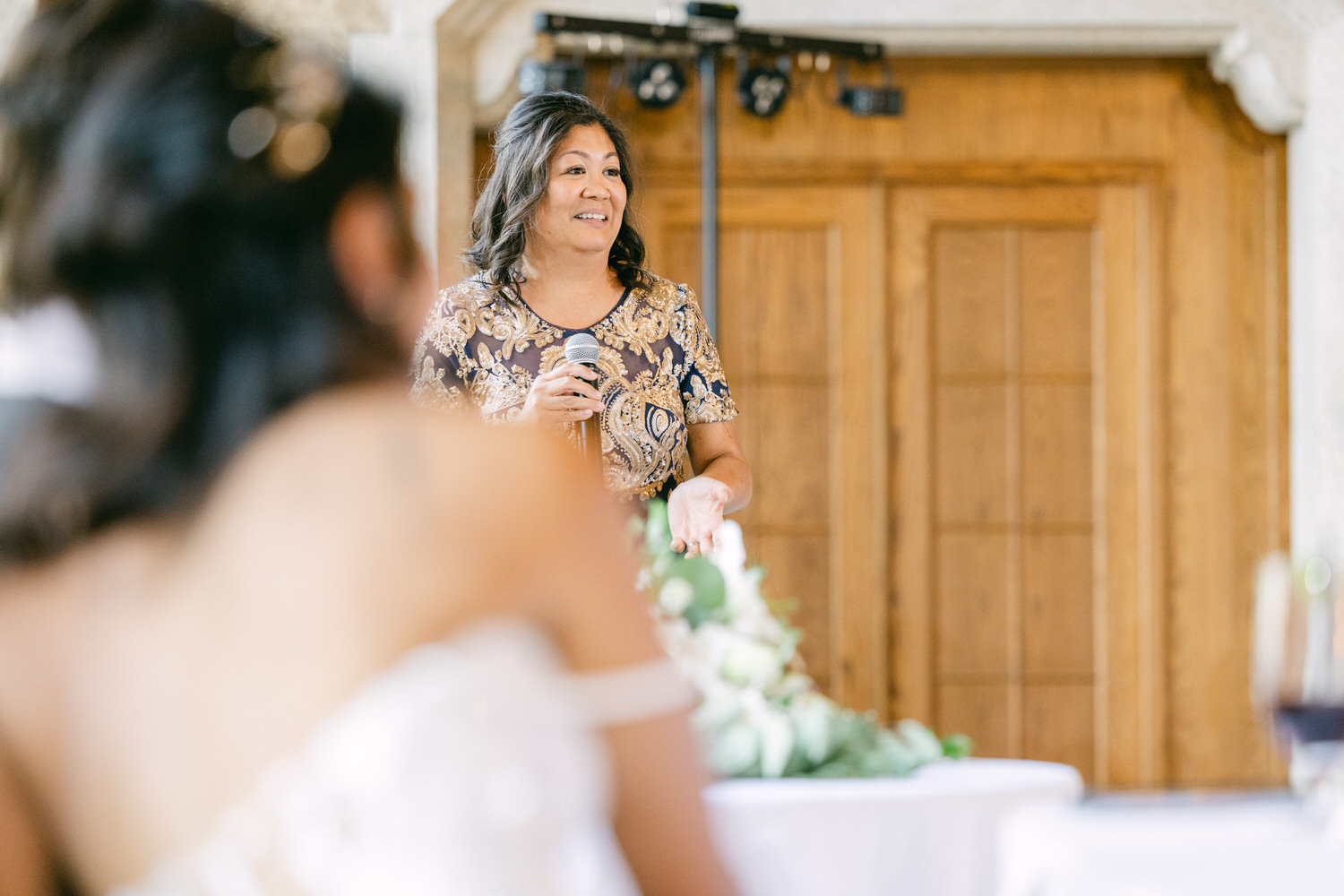 A woman delivers a heartfelt speech at a wedding reception, with guests attentively listening in the foreground.