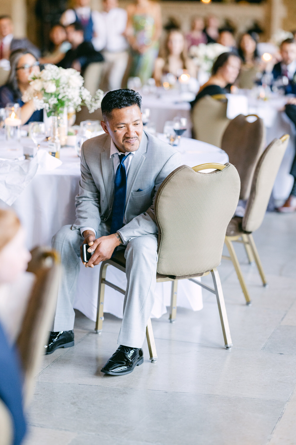 A well-dressed man in a light gray suit and blue tie sits on a chair at a wedding reception, observing the gathering with a smile, surrounded by elegantly set tables and guests in the background.