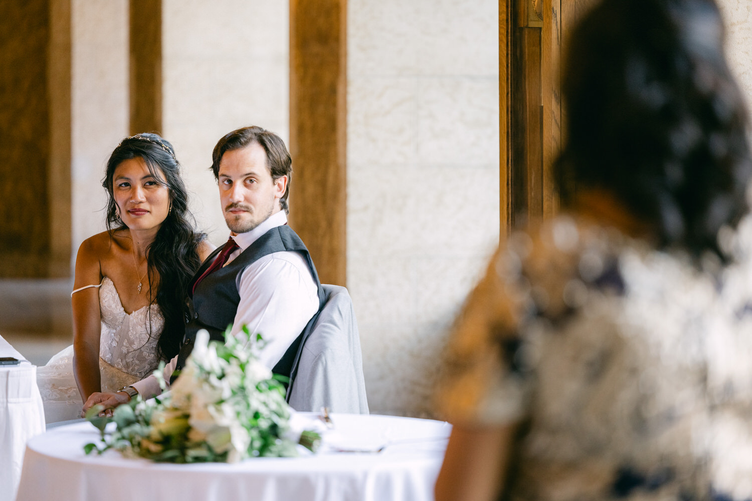 A couple sitting at a table adorned with flowers, gazing at a guest in a beautifully lit indoor setting.