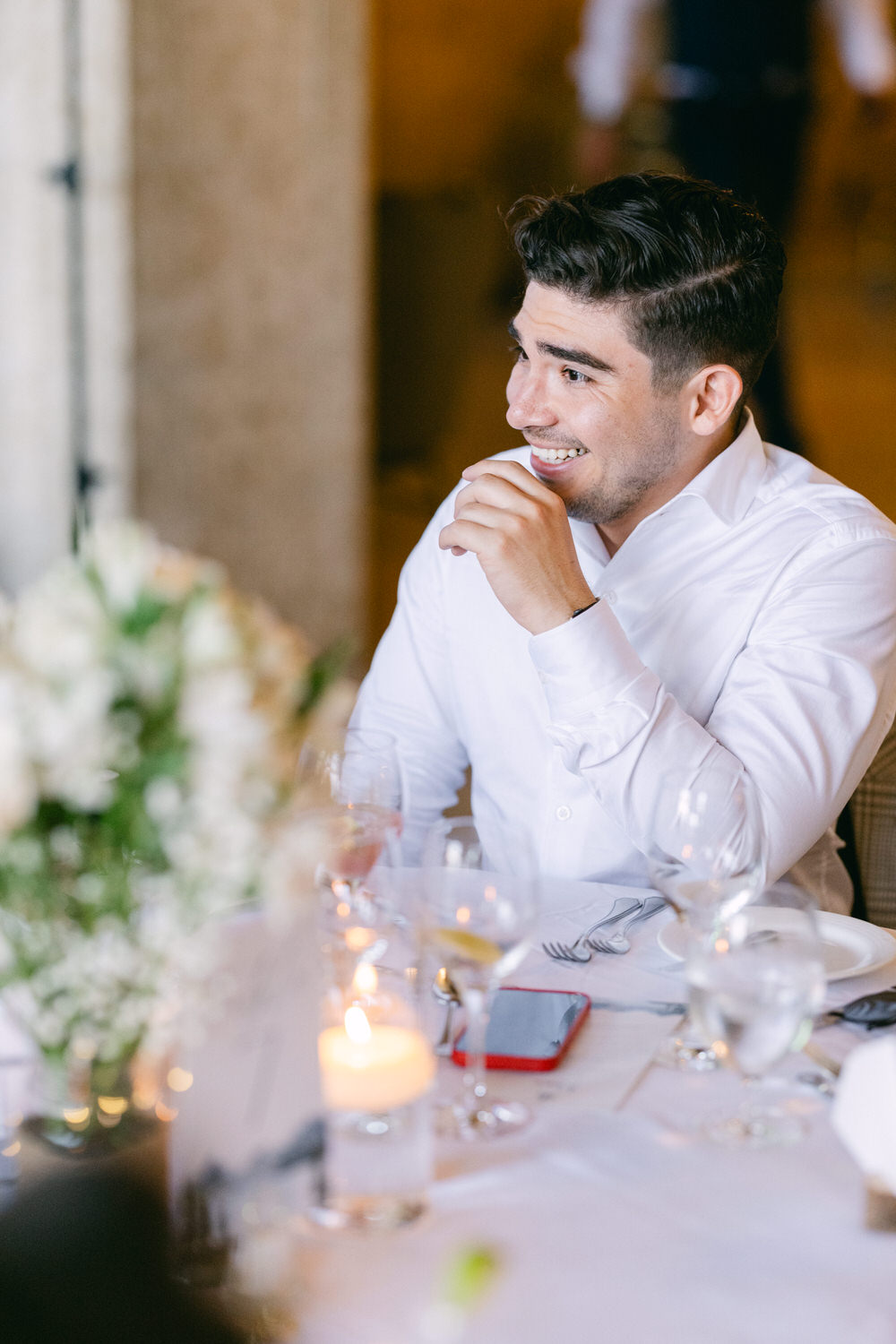 A young man in a white shirt smiles and engages in conversation at a dining table adorned with flowers and dinnerware.