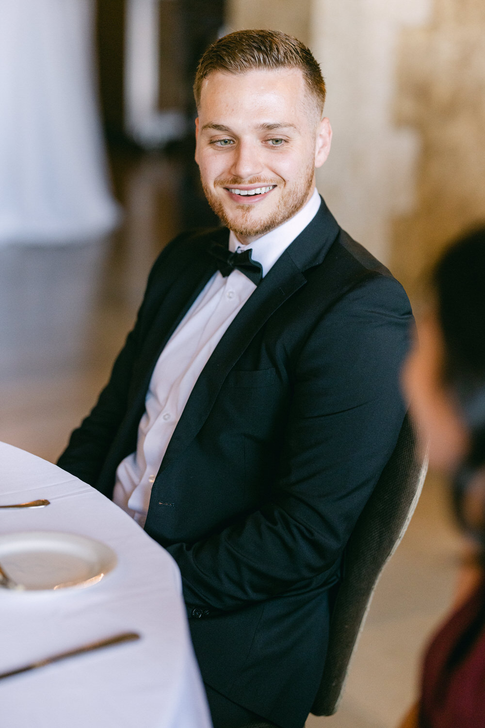 A well-dressed man in a tuxedo smiles while seated at a table, featuring a white tablecloth and a plate in front of him.