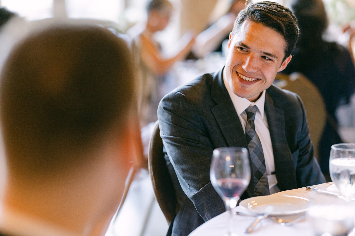 A well-dressed man smiling and engaging in conversation at a dining table during a social gathering, with glasses of wine and water on the table.