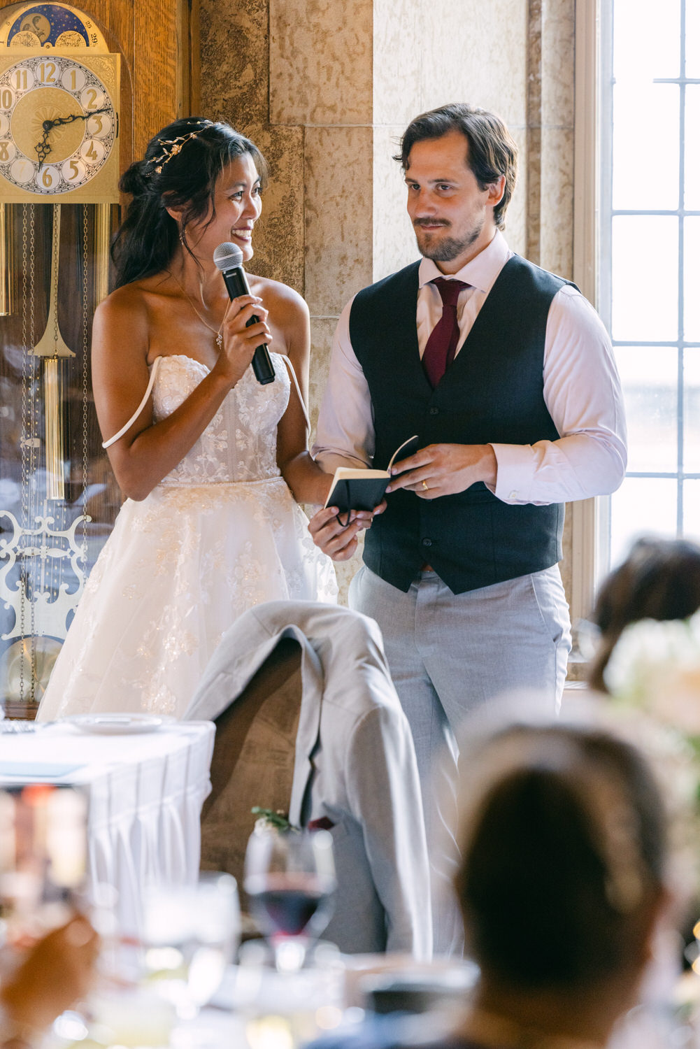 A bride and groom share a heartfelt moment as the bride speaks into a microphone while the groom holds a notepad, with guests watching in a beautifully decorated venue.