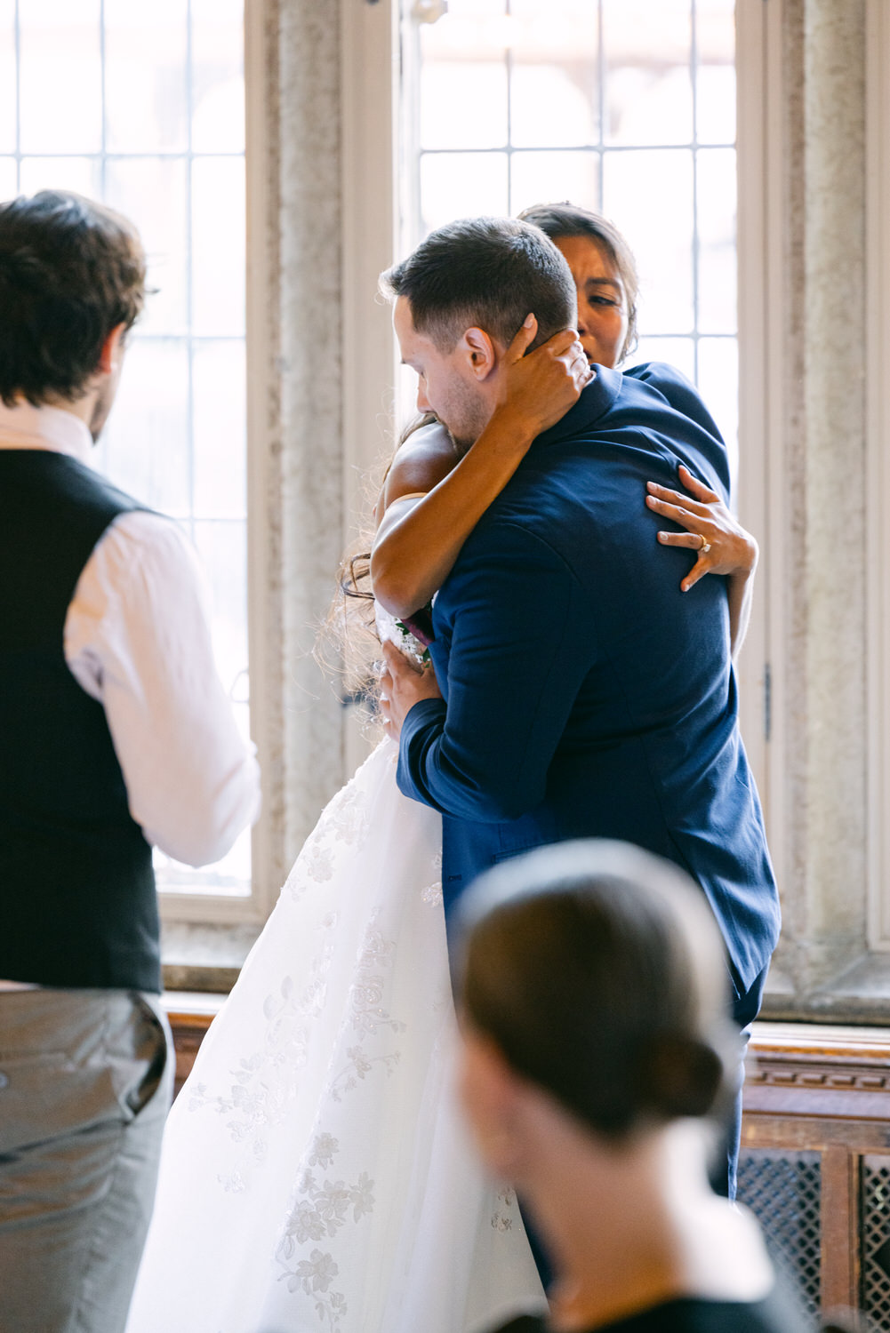 A joyful couple shares a heartfelt hug during their wedding ceremony, surrounded by guests in a beautifully lit room.