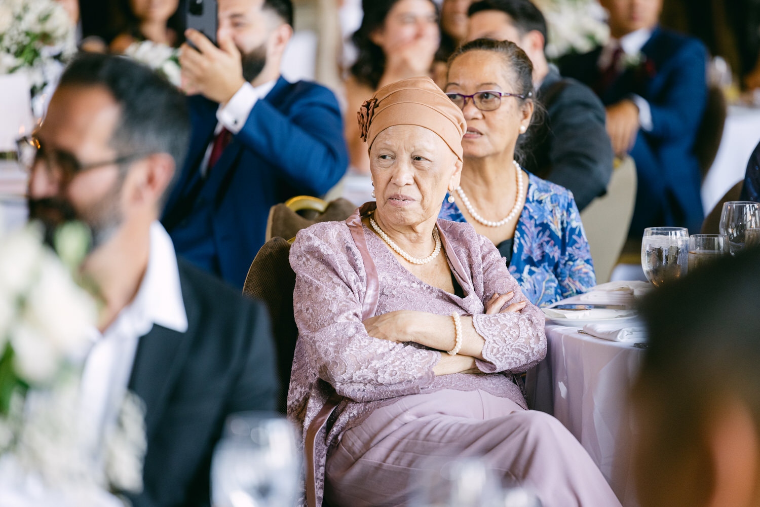 An elderly woman with a headscarf and a delicate purple outfit sits at a wedding reception, looking thoughtfully at the event, surrounded by other guests in a festive setting.