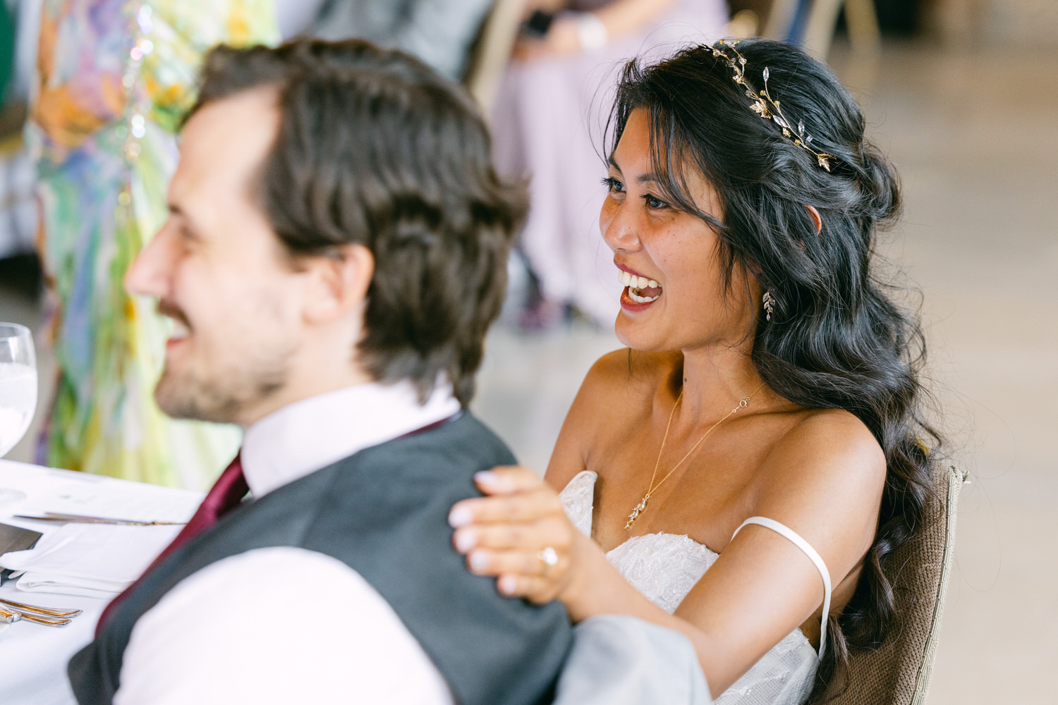 A joyful bride smiles while seated next to a groom at a wedding reception, capturing a moment of laughter and celebration.